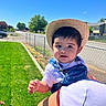 toddler, child, hat, straw_hat, denim_overalls, outdoor, sunny, daytime, grass, blue_sky, person, holding, fence, sidewalk, residential_area, casual_clothing, drool, curious_expression, portrait, baby
