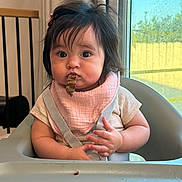 Kendall is registered to the contest to win money with this photo: baby, child, high_chair, bib, food, indoor, curtains, window, hair_clip, messy, face, mouth, hands, table, seat, curious, skin, clothing, person, infant
