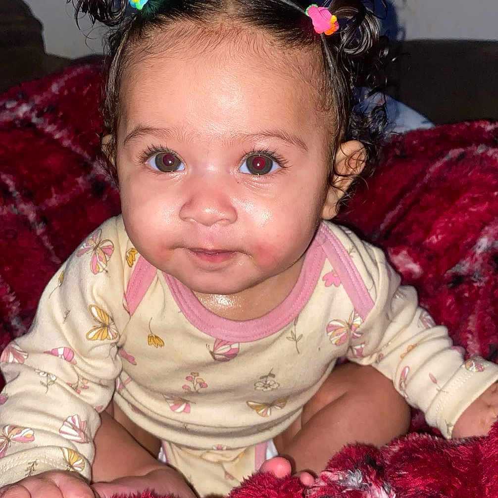 Solana is registered to the contest to win money with this photo: baby, child, infant, cute, hair_ties, onesie, blanket, red_blanket, sitting, indoors, face, portrait, young_child, curious, bright_eyes, smile, skin, hand, fingers, cozy