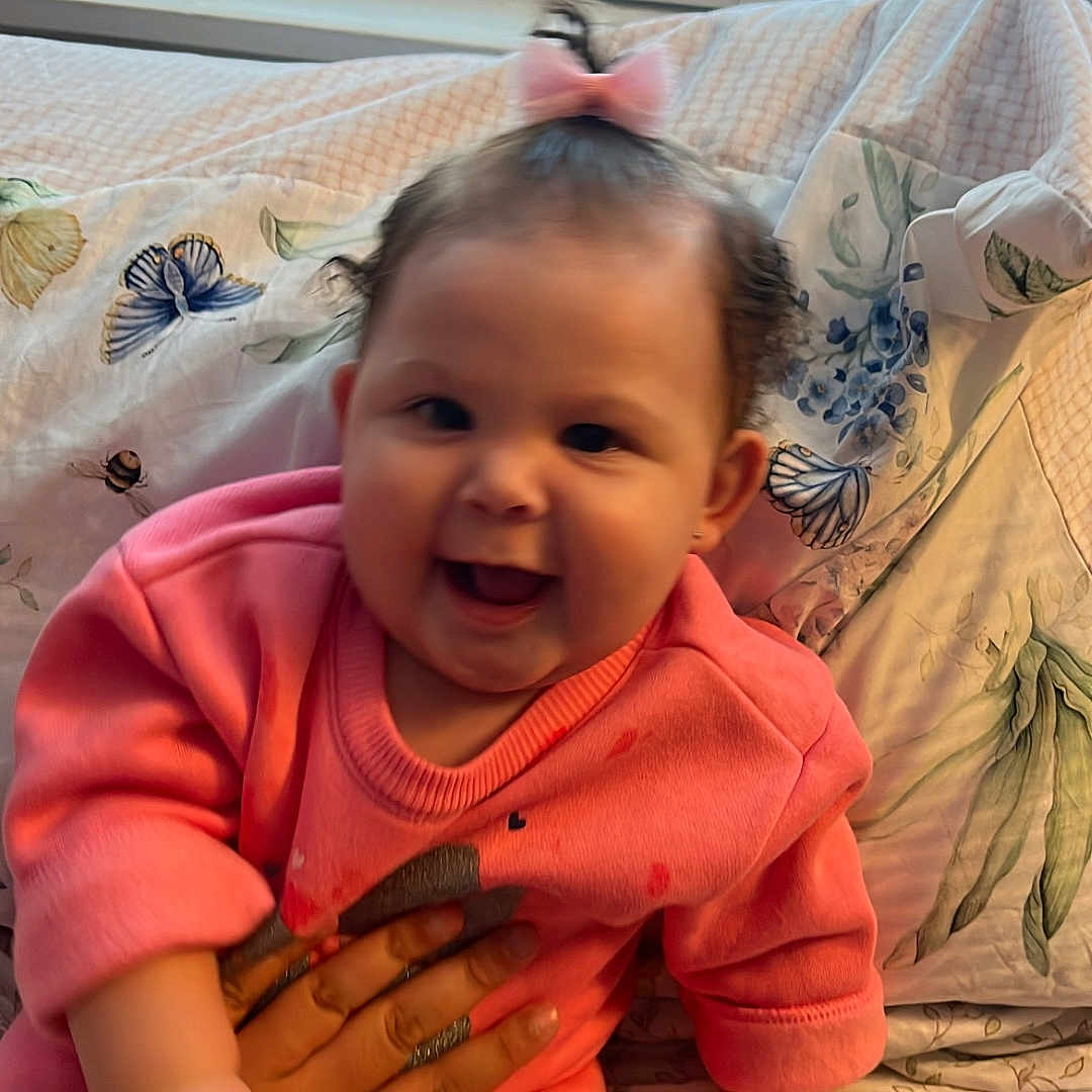 Yma Isabell is registered to the contest to win money with this photo: baby, bedspread, bow, child, clothing, cute, face, fingers, floral_pattern, hand, happy, indoors, infant, person, pink_outfit, portrait, sitting, skin, smiling, window_blinds