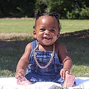 Major is registered to the contest to win money with this photo: baby, child, denim_overalls, gold_chain, smiling, happy, outdoor, grass, blanket, cute, portrait, infant, sitting, sunlight, greenery, young_child, face, skin, feet, hair