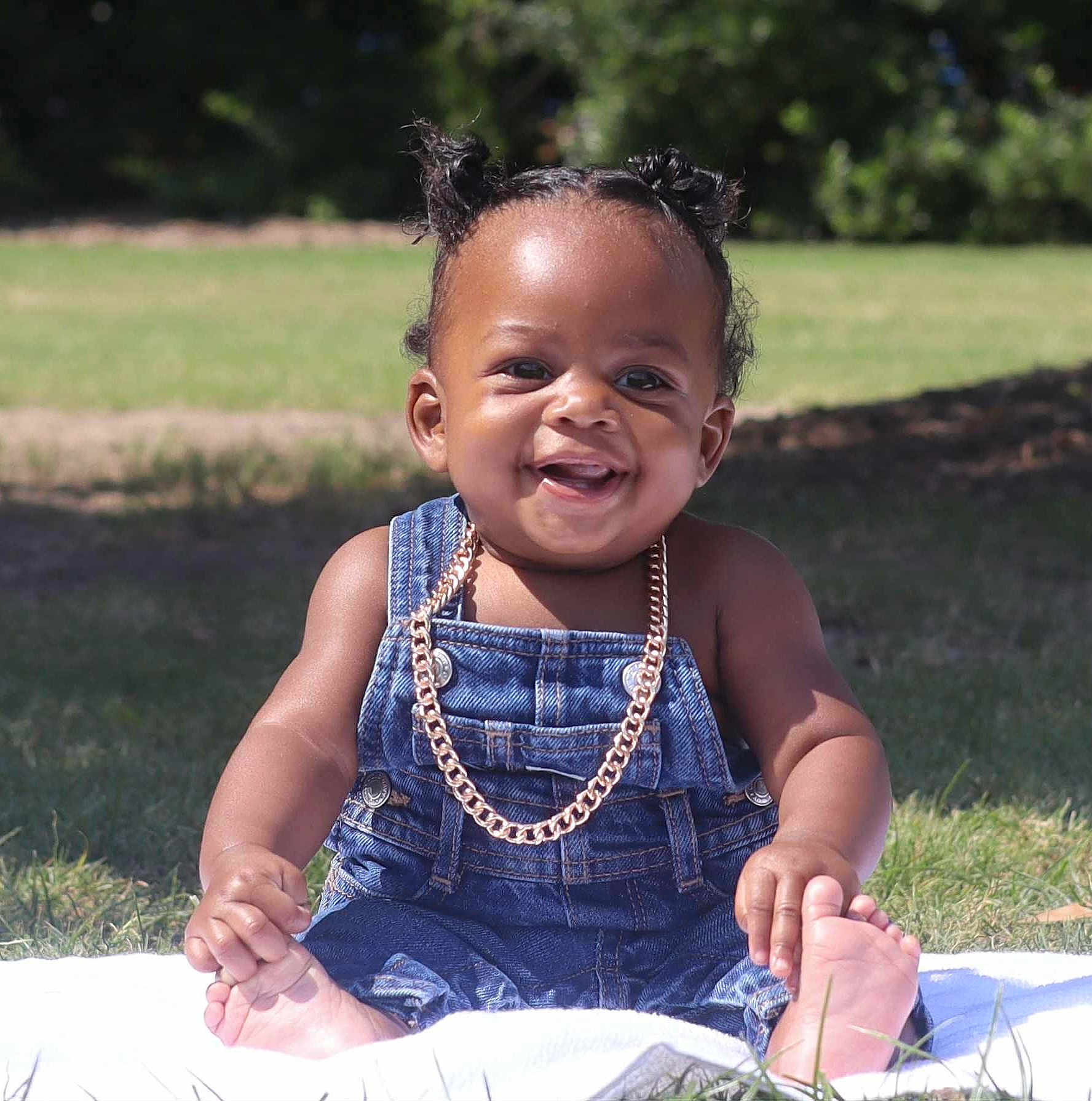 Major is registered to the contest to win money with this photo: baby, child, denim_overalls, gold_chain, smiling, happy, outdoor, grass, blanket, cute, portrait, infant, sitting, sunlight, greenery, young_child, face, skin, feet, hair