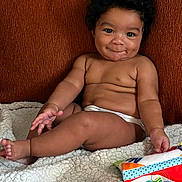 Naomi joined the competition — help win amazing prizes! baby, infant, smiling, sitting, blanket, toy, couch, indoor, child, cute, playful, diaper, cozy, happy, face, person, curly_hair, skin, comfort, relaxing