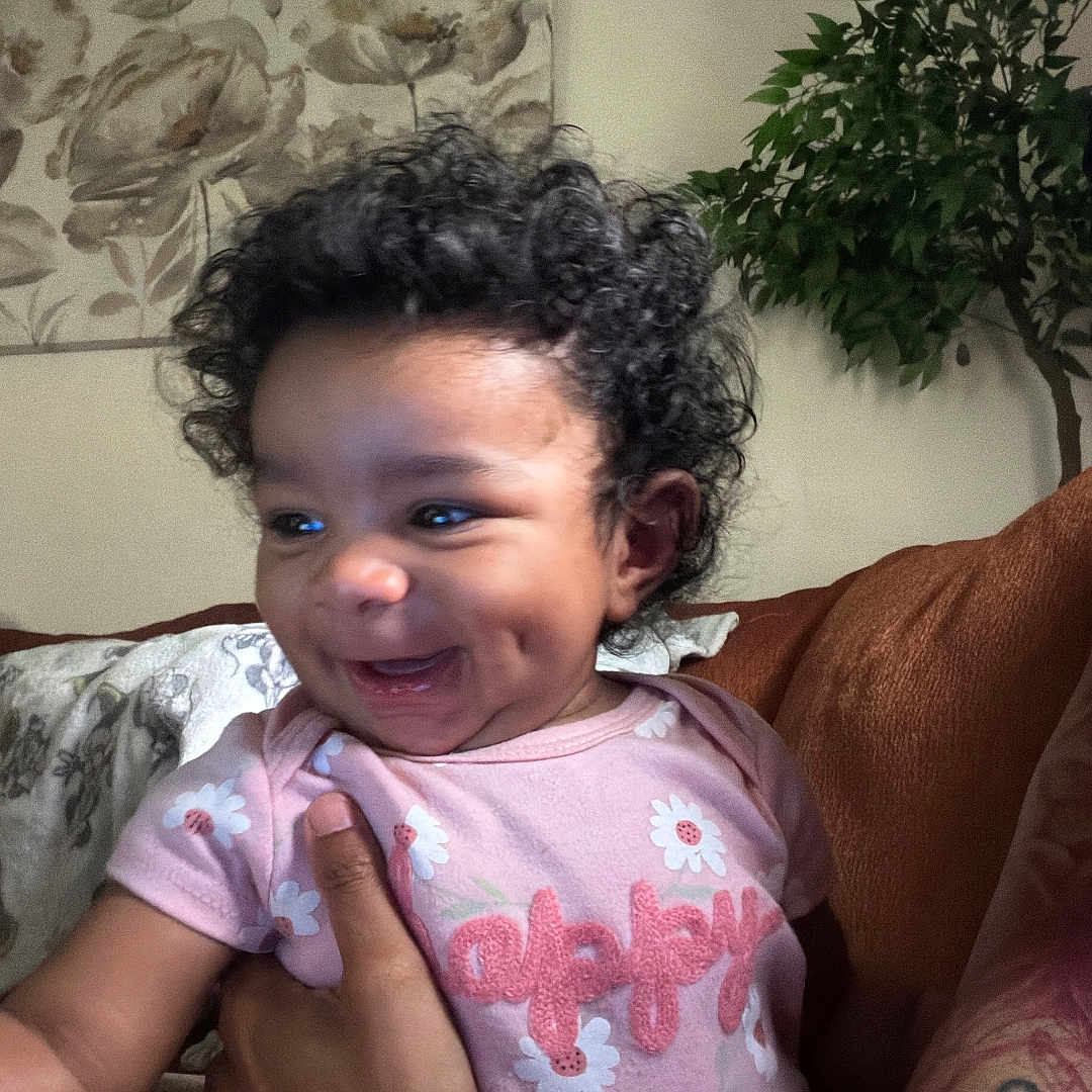 Naomi is registered to the contest to win money with this photo: baby, child, curly_hair, pink_onesie, smiling, dimples, tattoo, arm, hand, plant, living_room, couch, floral_art, indoor, person, happy, portrait, family, holding, cute