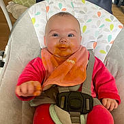 Maëlya participe au concours pour gagner de l'argent avec cette photo : baby, high_chair, bib, baby_food, messy, pink_clothing, smile, face, hands, indoor, feeding, child, happy, seatbelt, wooden_floor, background, cushion, person, infant, home