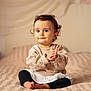 baby, bed, black_pants, blanket, child, clapping, cozy, curly_hair, cute, expression, face, indoors, infant, long_sleeve_shirt, person, sitting, smiling, soft_lighting, warm_colors, young
