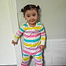 child, toddler, girl, portrait, standing, smiling, pigtails, hair_bows, striped_clothing, rainbow_colors, onesie, pajamas, indoors, door, vent, carpet, home, full_body, happy, fashion