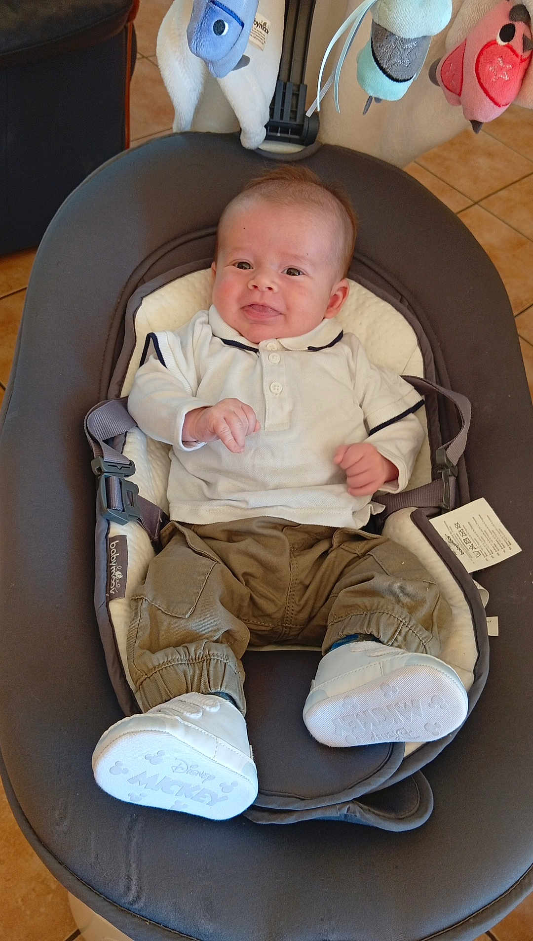 Ayden a rejoint le concours — aidez-le/la à gagner de superbes lots ! baby, infant, baby_rocker, white_shirt, khaki_pants, mickey_mouse_shoes, soft_toys, indoor, smiling, cute, comfortable, child, footwear, seat, baby_clothing, floor_tiles, playful, happy, person, young_child