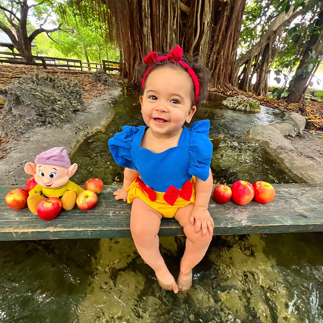 Yailee joined the competition — help win amazing prizes! apples, bench, blue_dress, child, costume, cute, daylight, feet_in_water, greenery, happy, nature, outdoor, park, playful, plush_toy, red_headband, smiling, toddler, tree_roots, water