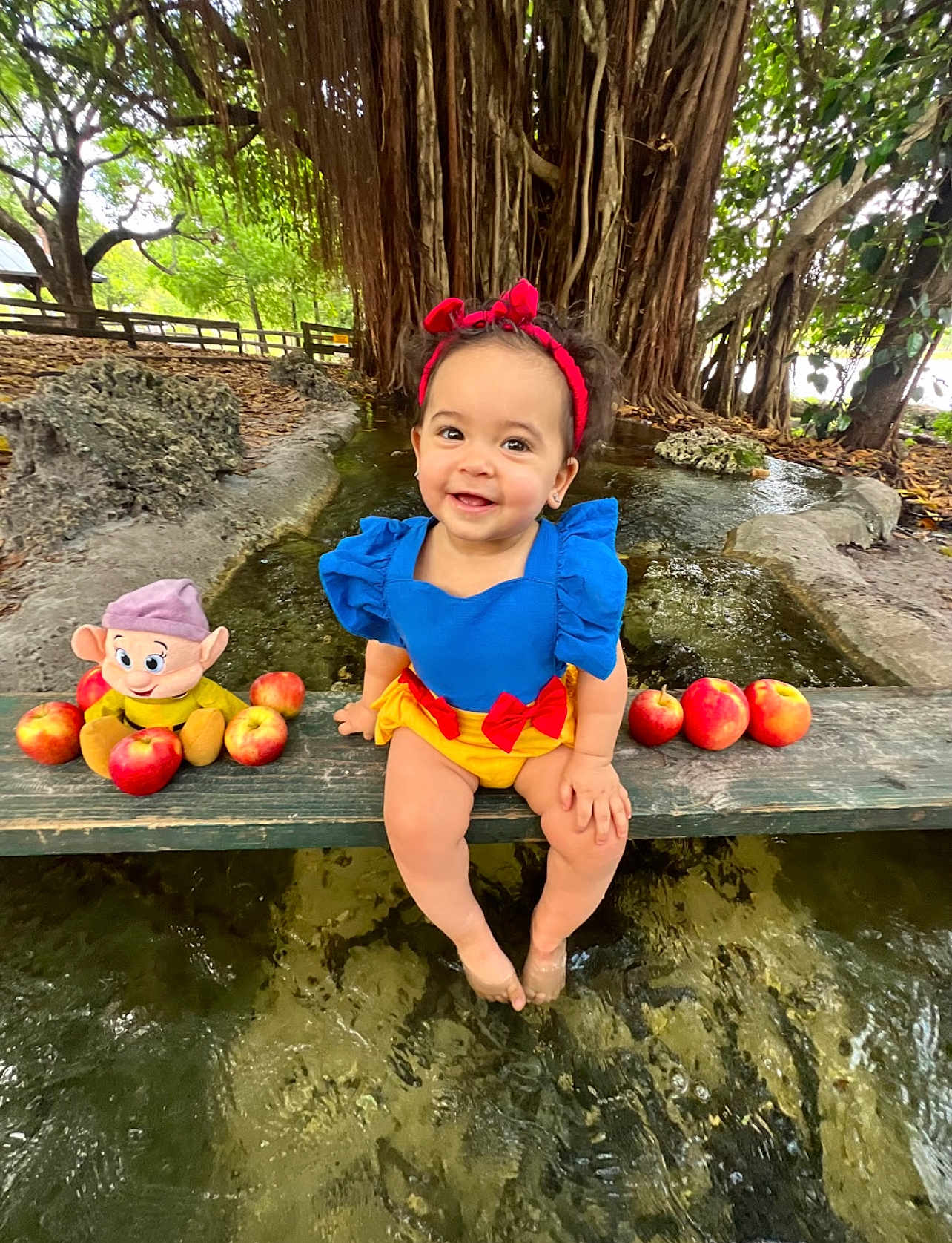 Yailee joined the competition — help win amazing prizes! toddler, child, smiling, blue_dress, red_headband, apples, plush_toy, water, tree_roots, outdoor, nature, bench, park, happy, feet_in_water, costume, playful, cute, daylight, greenery