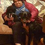 Bear is registered to the contest to win money with this photo: person, dog, small_dog, large_dog, couch, window, blind, pillow, snowman, floor, shoe, indoor, casual_clothing, sitting, pet, home, cozy, black_dog, brown_dog, happy