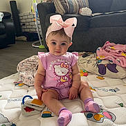Lillith is registered to the contest to win money with this photo: baby, child, infant, sitting, portrait, pink_bow, hair_accessory, hello_kitty, pink_outfit, socks, smiling, blue_eyes, indoors, living_room, couch, blanket, quilt, toys, pillows, home