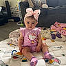 baby, blanket, blue_eyes, child, couch, hair_accessory, hello_kitty, home, indoors, infant, living_room, pillows, pink_bow, pink_outfit, portrait, quilt, sitting, smiling, socks, toys