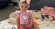 Lillith is registered to the contest to win money with this photo: baby, child, infant, portrait, sitting, indoors, bedroom, quilt, blanket, pink_bow, hair_accessory, hello_kitty, pajamas, socks, smiling, face, toy, couch, soft_lighting, home