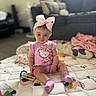 baby, bedroom, blanket, child, couch, face, hair_accessory, hello_kitty, home, indoors, infant, pajamas, pink_bow, portrait, quilt, sitting, smiling, socks, soft_lighting, toy