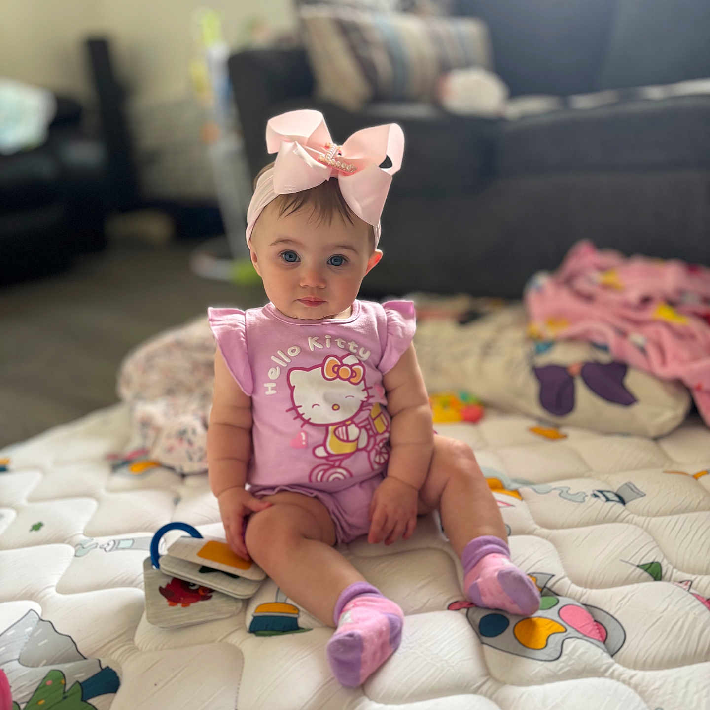 Lillith is registered to the contest to win money with this photo: baby, child, infant, portrait, sitting, indoors, bedroom, quilt, blanket, pink_bow, hair_accessory, hello_kitty, pajamas, socks, smiling, face, toy, couch, soft_lighting, home