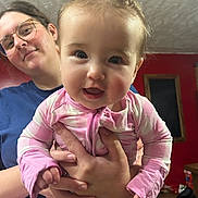 Avery is registered to the contest to win money with this photo: baby, child, woman, glasses, indoor, smile, happy, person, holding, pink_clothing, pigtails, face, portrait, young_child, cheerful, casual, home, fingers, hands, background