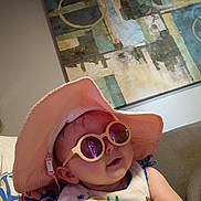 Natalie joined the competition — help win amazing prizes! baby, sunglasses, hat, indoor, bib, smiling, child, face, person, colorful, fashion, cute, portrait, accessory, happy, clothing, sitting, hand, couch, artwork