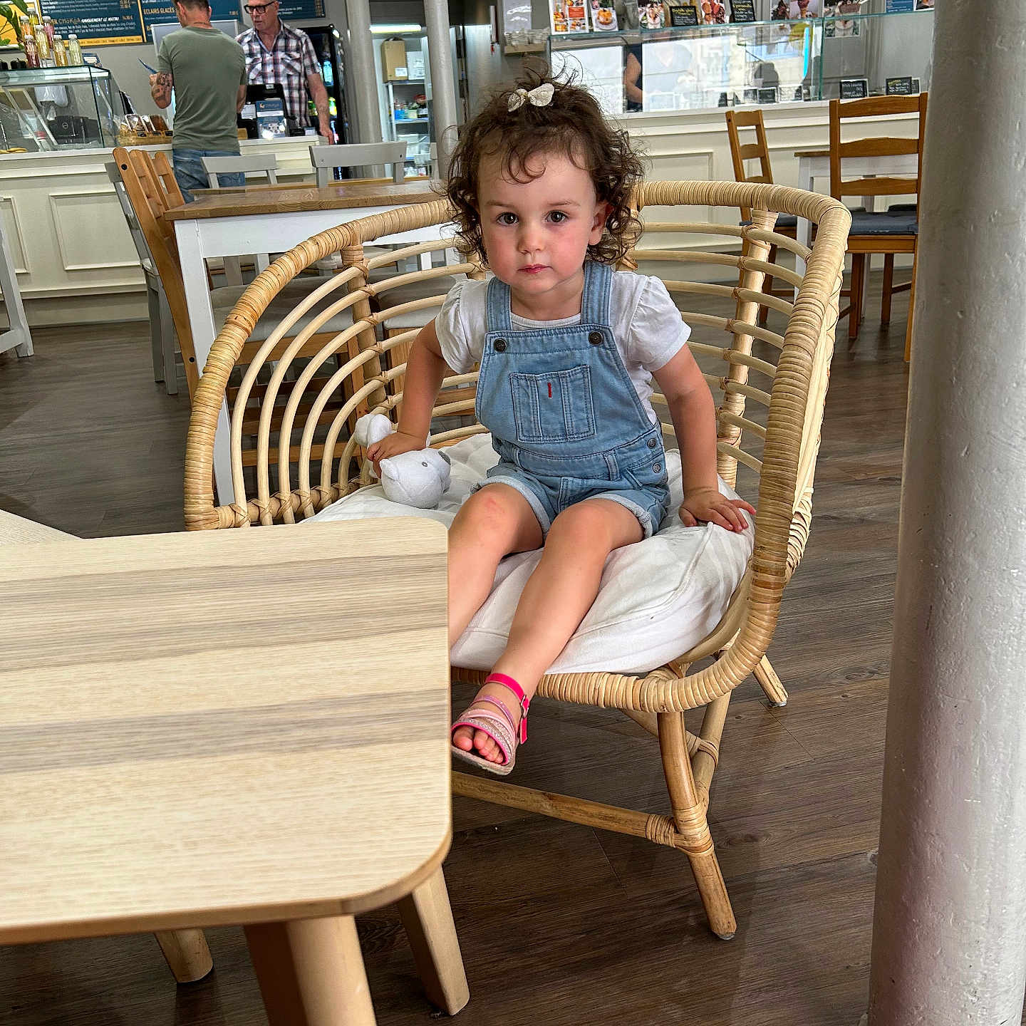 Erynn a rejoint le concours — aidez-le/la à gagner de superbes lots ! backpack, cafe, casual_clothing, child, counter, curly_hair, denim_overalls, flooring, furniture, indoor, menu, people, pillar, pink_sandals, serious_expression, stuffed_toy, table, toddler, wicker_chair, wooden_floor