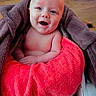 baby, smiling, towel, blanket, happy, infant, face, skin, wood_floor, wrapped, cozy, child, indoors, portrait, cute, hands, head, bright_color, expression, young