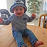 baby, child, striped_clothing, hat, pirate_hat, barefoot, wooden_table, indoor, plant, stroller, curtains, smiling, happy, jeans, furniture, person, face, sitting, natural_light, home