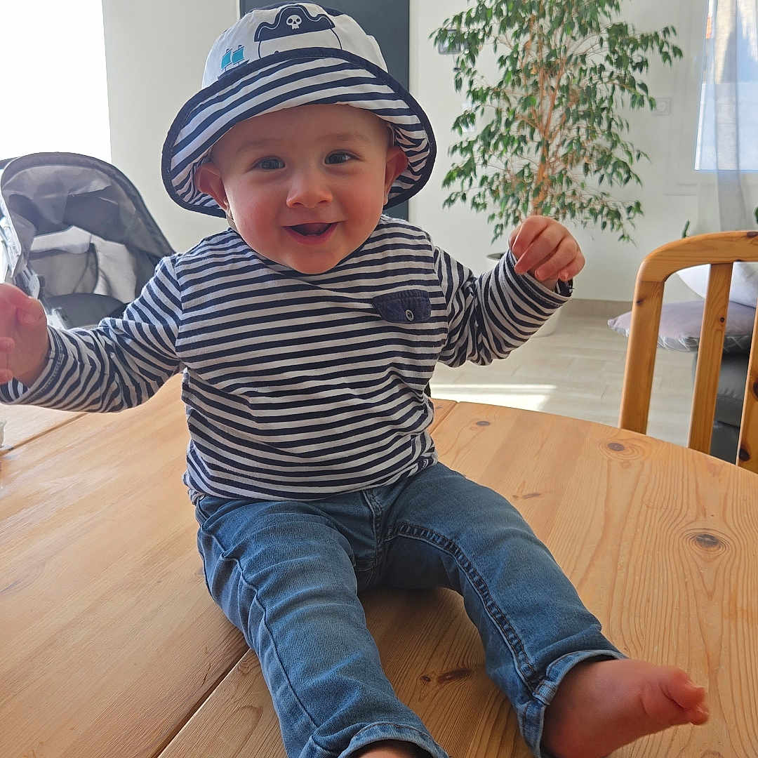 Tymaël participe au concours pour gagner de l'argent avec cette photo : baby, child, striped_clothing, hat, pirate_hat, barefoot, wooden_table, indoor, plant, stroller, curtains, smiling, happy, jeans, furniture, person, face, sitting, natural_light, home