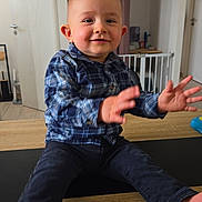 Tymaël participe au concours pour gagner de l'argent avec cette photo : toddler, child, smiling, blue_shirt, plaid, indoor, table, wooden_table, baby_gate, hands, feet, pants, happy, person, home, curtain, door, floor, furniture, playful