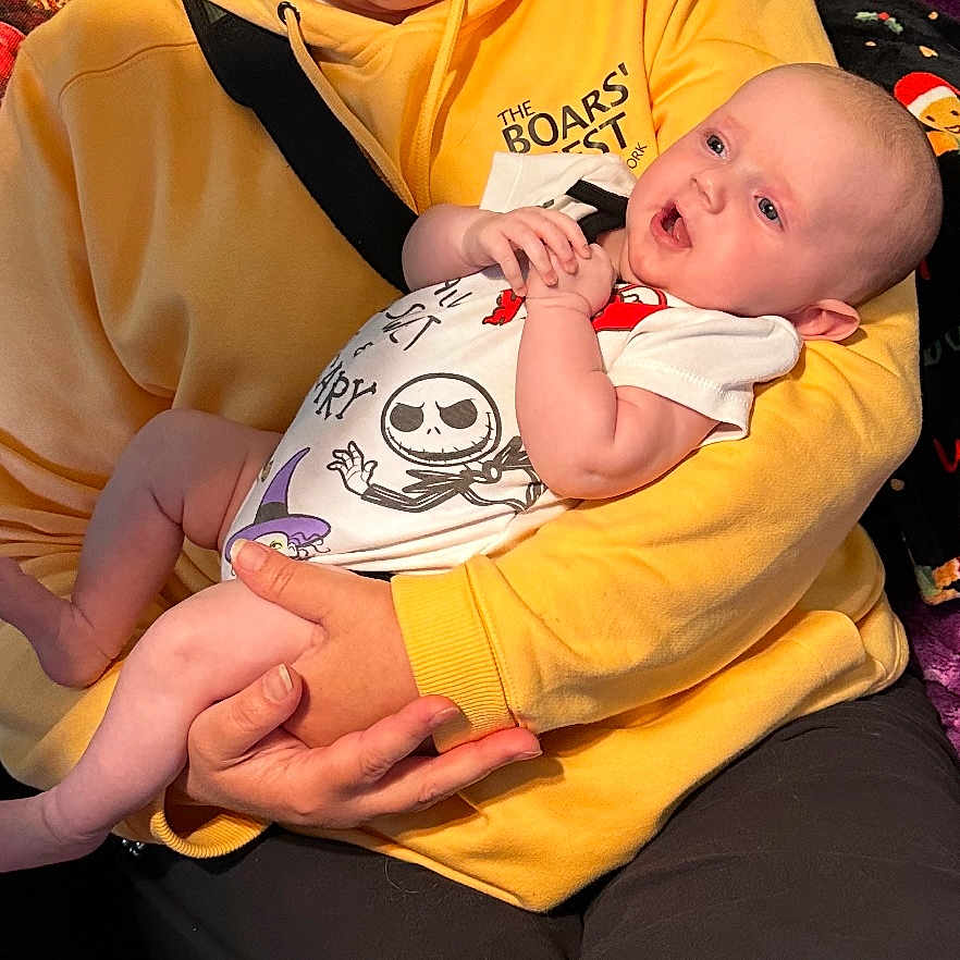 Saraya is registered to the contest to win money with this photo: arm, baby, black_pants, blanket, closeup, clothing, cute, face, festive, hand, holding, holiday, indoor, infant, onesie, person, purple_blanket, relaxed, smiling, yellow_hoodie