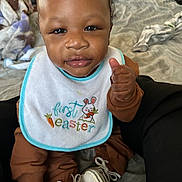 Ares joined the competition — help win amazing prizes! baby, child, infant, bib, first_easter, thumbs_up, brown_clothing, white_shoes, indoor, blanket, bed, floor, person, cute, portrait, young_child, sitting, expression, cozy, soft_light