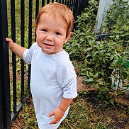 John is registered to the contest to win money with this photo: toddler, child, baby, portrait, smiling, outdoors, garden, fence, backyard, plants, grass, white_outfit, blue_shoes, standing, walking, red_hair, face, person, daylight, photography
