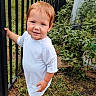 baby, backyard, blue_shoes, child, daylight, face, fence, garden, grass, outdoors, person, photography, plants, portrait, red_hair, smiling, standing, toddler, walking, white_outfit