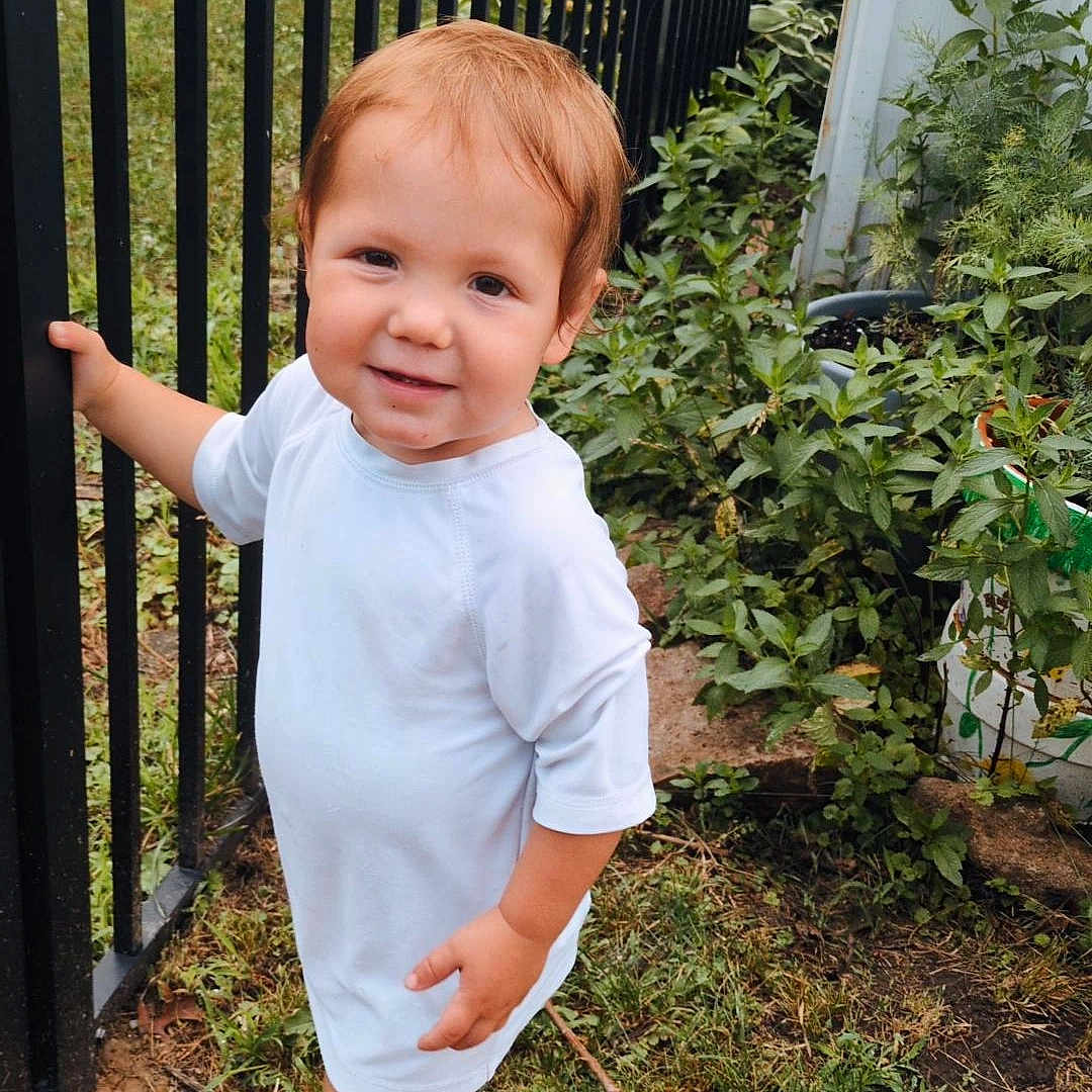 John is registered to the contest to win money with this photo: toddler, child, baby, portrait, smiling, outdoors, garden, fence, backyard, plants, grass, white_outfit, blue_shoes, standing, walking, red_hair, face, person, daylight, photography