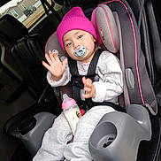 Victoria is registered to the contest to win money with this photo: toddler, child, car_seat, pacifier, bottle, hat, beanie, gray_clothing, car_interior, seatbelt, cup_holder, window, houses, hand_raised, footwear, sneakers, baby, person, seat, vehicle