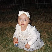 Victoria is registered to the contest to win money with this photo: child, toddler, girl, white_clothing, headband, flashlight, grass, night, outdoor, wire_fence, curious, crouching, polka_dot, long_sleeve, dress, headwear, person, young_child, looking_up, dark_background