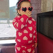 Victoria is registered to the contest to win money with this photo: brick_wall, casual_wear, child, cozy, cute, fashion, fun, headband, heart_pattern, heart_sunglasses, indoor, person, playful, portrait, red_clothing, smiling, standing, toddler, window, young_child