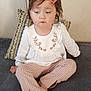 child, girl, bow, pink_bow, white_top, embroidered_clothing, patterned_pants, sitting, couch, cushion, indoor, person, foot, long_sleeves, wall, home, portrait, casual, young_child, expression