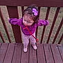 toddler, child, purple_sweater, bow, red_ribbon, wooden_deck, wooden_railing, grass, standing, pink_shoes, patterned_pants, outdoor, person, young_child, curly_hair, playful, casual_clothing, daylight, nature, balcony
