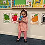 toddler, child, indoor, carpet, pink_dress, leggings, sippy_cup, educational_posters, color_posters, frog, apple, pumpkin, black_shoes, bow, wall, learning, cute, standing, person, young_child