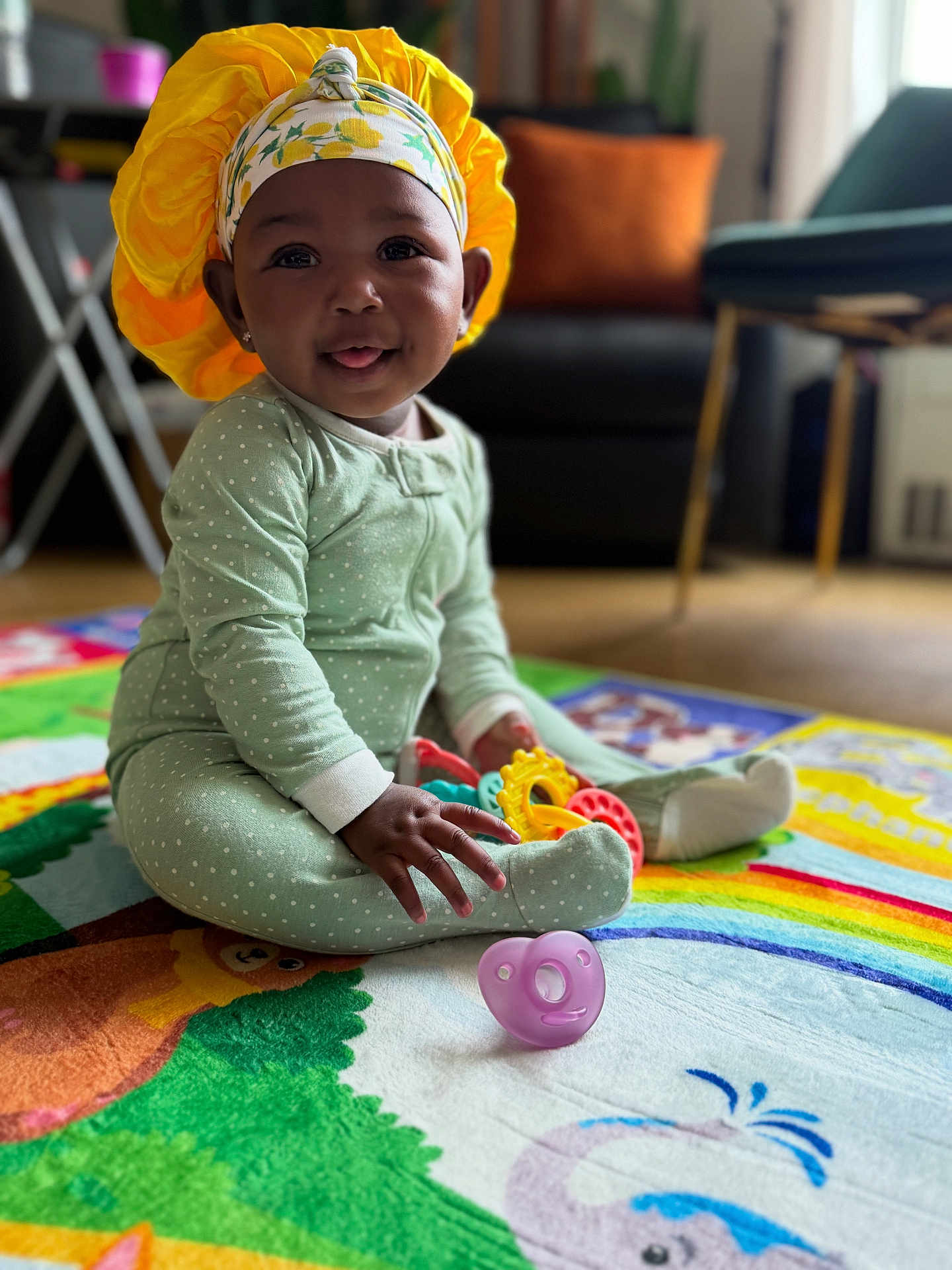 Nyla joined the competition — help win amazing prizes! baby, child, playmat, toys, onesie, headwear, yellow_bonnet, smiling, tongue_out, indoor, living_room, couch, colorful, floor, sitting, happy, cute, polka_dot, infant, playtime