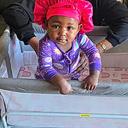 Nyla joined the competition — help win amazing prizes! baby, adult, bonnet, purple_onesie, playpen, indoor, floor, leather_couch, mirror, supporting_hand, smiling, child, person, furniture, home, cozy, flooring, patterned_clothing, holding, infant