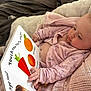 Mabel joined the competition — help win amazing prizes! baby, infant, pink_clothing, blanket, book, reading, child, board_book, orange_food, bed, cozy, knitted_blanket, hand, face, indoor, curious, lying_down, soft, cute, toddler