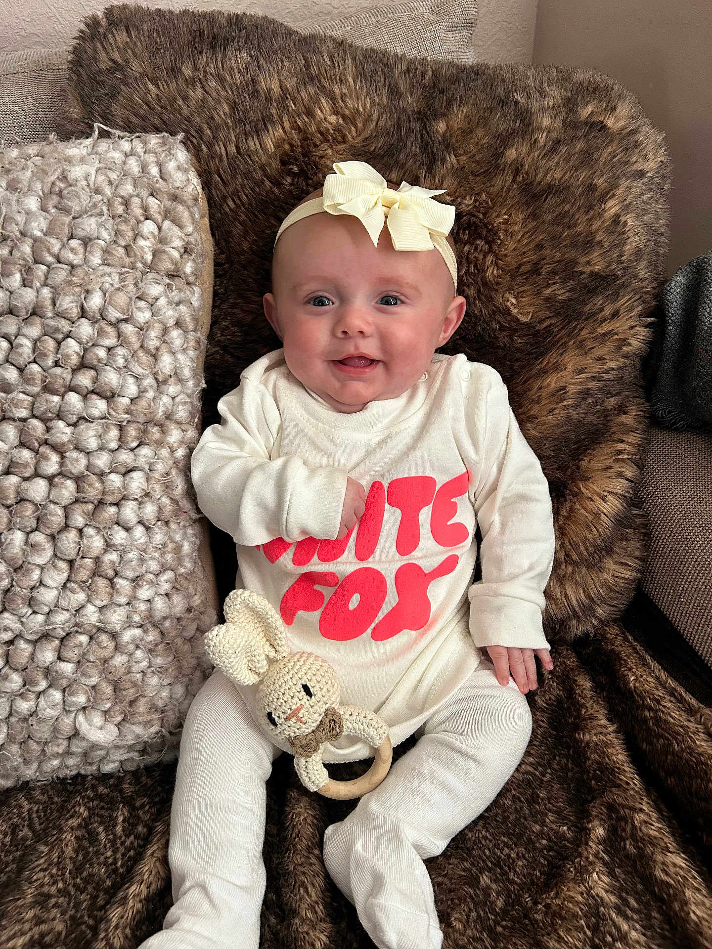 Mabel is registered to the contest to win money with this photo: baby, infant, smiling, headband, bow, white_clothing, text_on_clothing, knitted_toy, bunny_toy, blanket, furry_blanket, couch, cozy, indoor, cute, happy, portrait, child, sitting, soft_texture