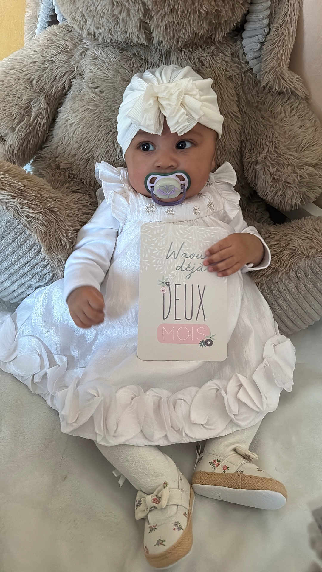 Ysalia participe au concours pour gagner de l'argent avec cette photo : baby, infant, pacifier, headwrap, bow, white_dress, milestone_card, teddy_bear, plush_toy, ruffles, floral_shoes, tights, sitting, portrait, closeup, cushion, soft_texture, indoor, hand, big_eyes