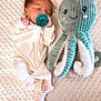 baby, newborn, pacifier, sleeping, plush_toy, octopus, stuffed_animal, blanket, soft_texture, cute, infant, child, cozy, white_clothing, striped_socks, bedtime, resting, smiling_toy, childhood, comfort