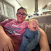 Eziah is registered to the contest to win money with this photo: baby, infant, woman, glasses, couch, sofa, living_room, t_shirt, smile, laughing, arm, hand, home_interior, kitchen, shelves, vent_hood, recessed_lighting, cozy, portrait, family
