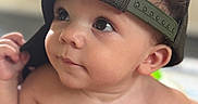 Zion joined the competition — help win amazing prizes! baby, infant, child, backwards_cap, hat, portrait, close_up, face, eyes, looking_up, curious, bare_chested, skin, adult_hand, finger, indoor, seated, red_blanket, soft_lighting, photography