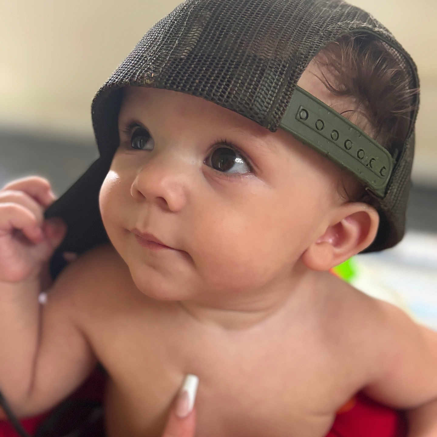 Zion joined the competition — help win amazing prizes! baby, infant, child, backwards_cap, hat, portrait, close_up, face, eyes, looking_up, curious, bare_chested, skin, adult_hand, finger, indoor, seated, red_blanket, soft_lighting, photography