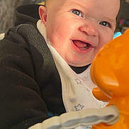 Leo joined the competition — help win amazing prizes! baby, smiling, toy, orange, black_clothing, bib, indoor, closeup, face, hand, child, infant, happy, cute, play, bright, light, expression, person, young