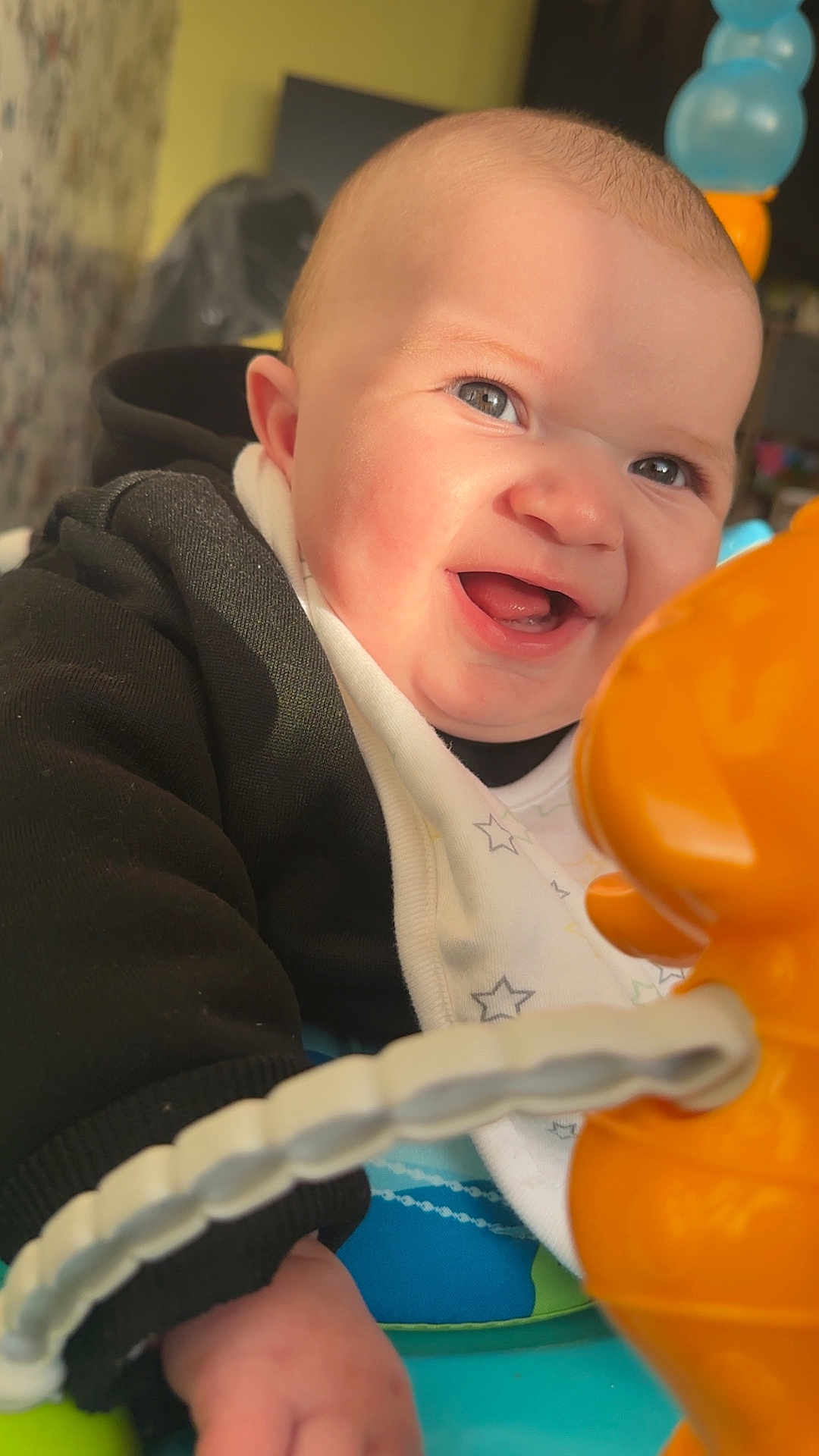 Leo joined the competition — help win amazing prizes! baby, smiling, toy, orange, black_clothing, bib, indoor, closeup, face, hand, child, infant, happy, cute, play, bright, light, expression, person, young