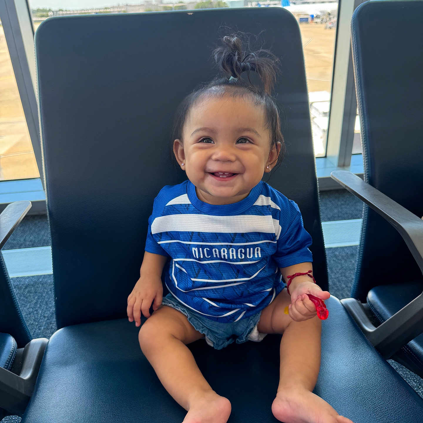 Sara Alina is registered to the contest to win money with this photo: airplane, airport, blue_shirt, casual_clothing, chair, child, cute, daylight, denim_shorts, footwear_free, happy, indoor, person, pony_tail, runway, sitting, smiling, toddler, waiting_area, window